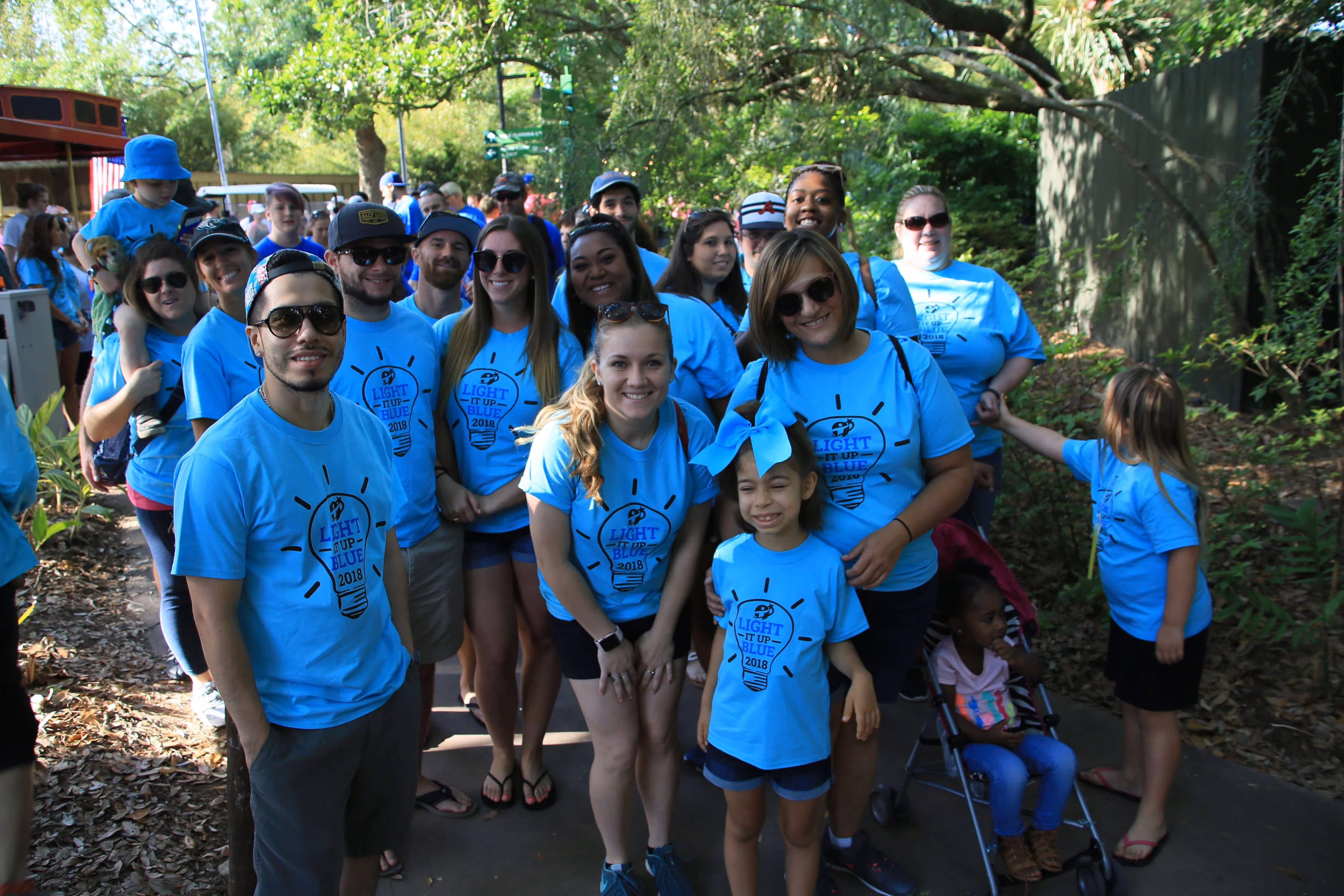 Group of people wearing matching blue 'Light It Up Blue 2018' t-shirts gathered outdoors in a shaded area with trees.