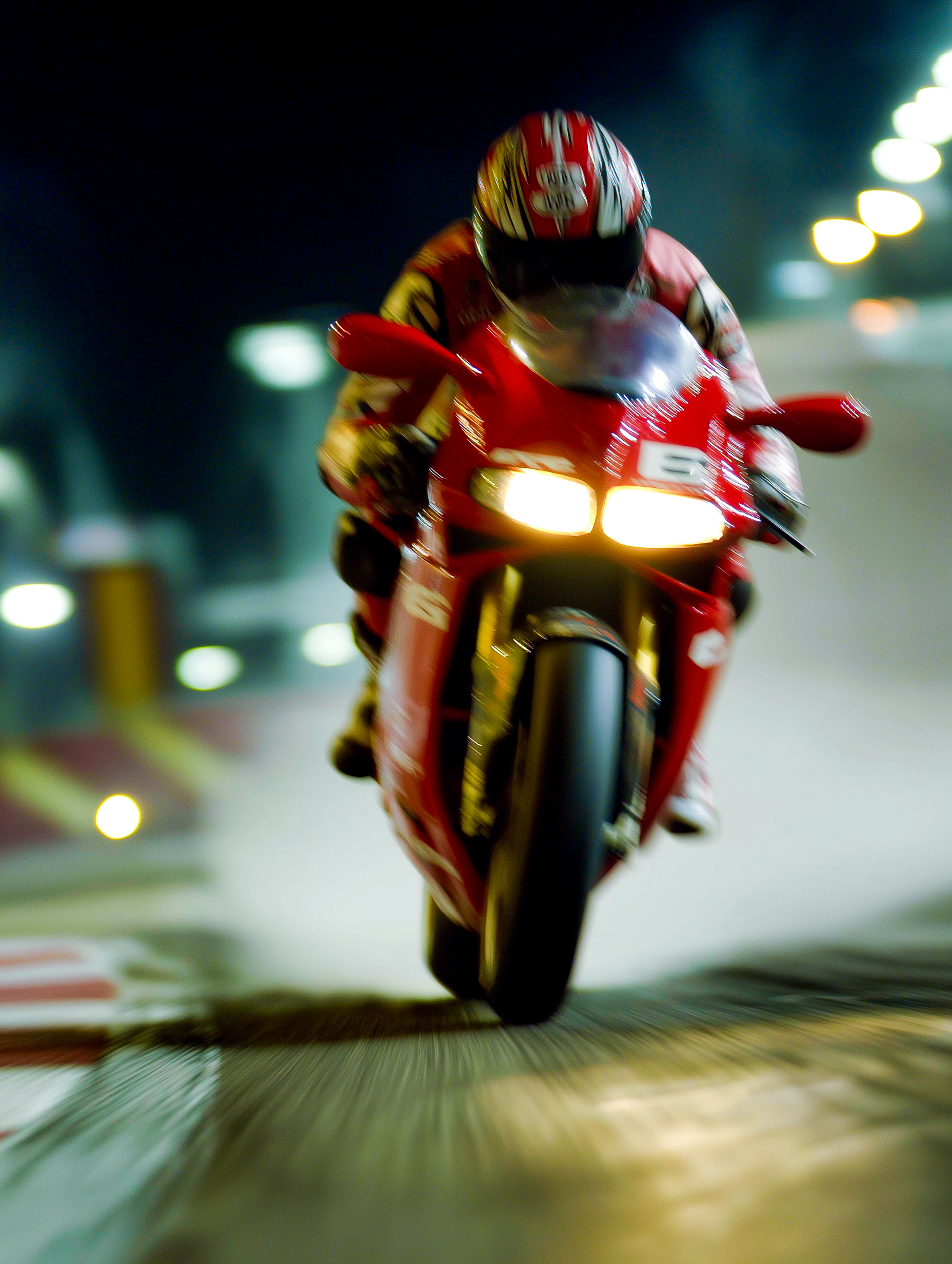 Motorcycle racer in red gear on a red sportbike racing on a track at night with headlights on.
