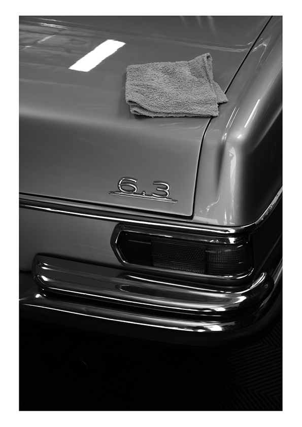 Close-up of a classic car rear corner showing the chrome bumper, tail light, and emblem with '6.3,' with a folded cloth on the trunk lid.