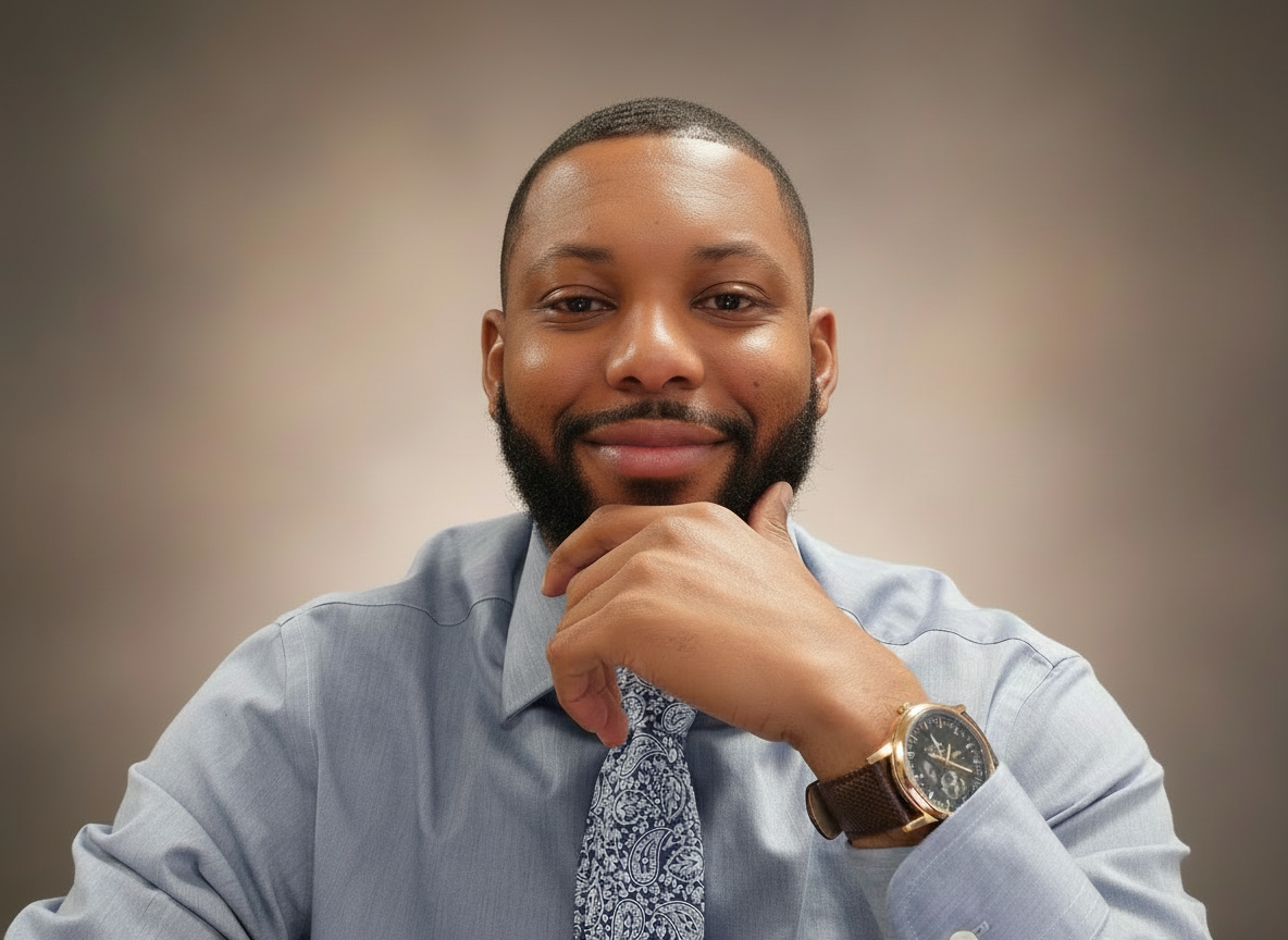 Smiling man with beard wearing a light blue dress shirt and paisley patterned tie, resting his chin on his hand, showing a wristwatch.