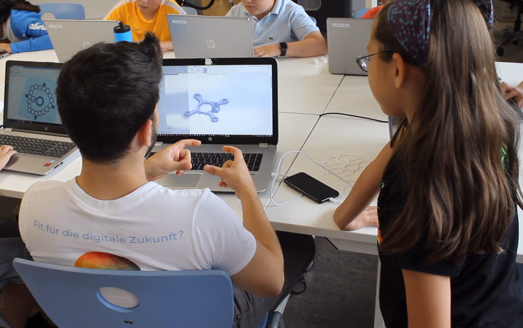 Ein Schüler zeigt einem Klassenkameraden in einem Klassenzimmer ein 3D-Modell auf einem Laptop, während andere Schüler im Hintergrund an Laptops arbeiten. Auf dem T-Shirt des Schülers steht "Fit für die digitale Zukunft?".