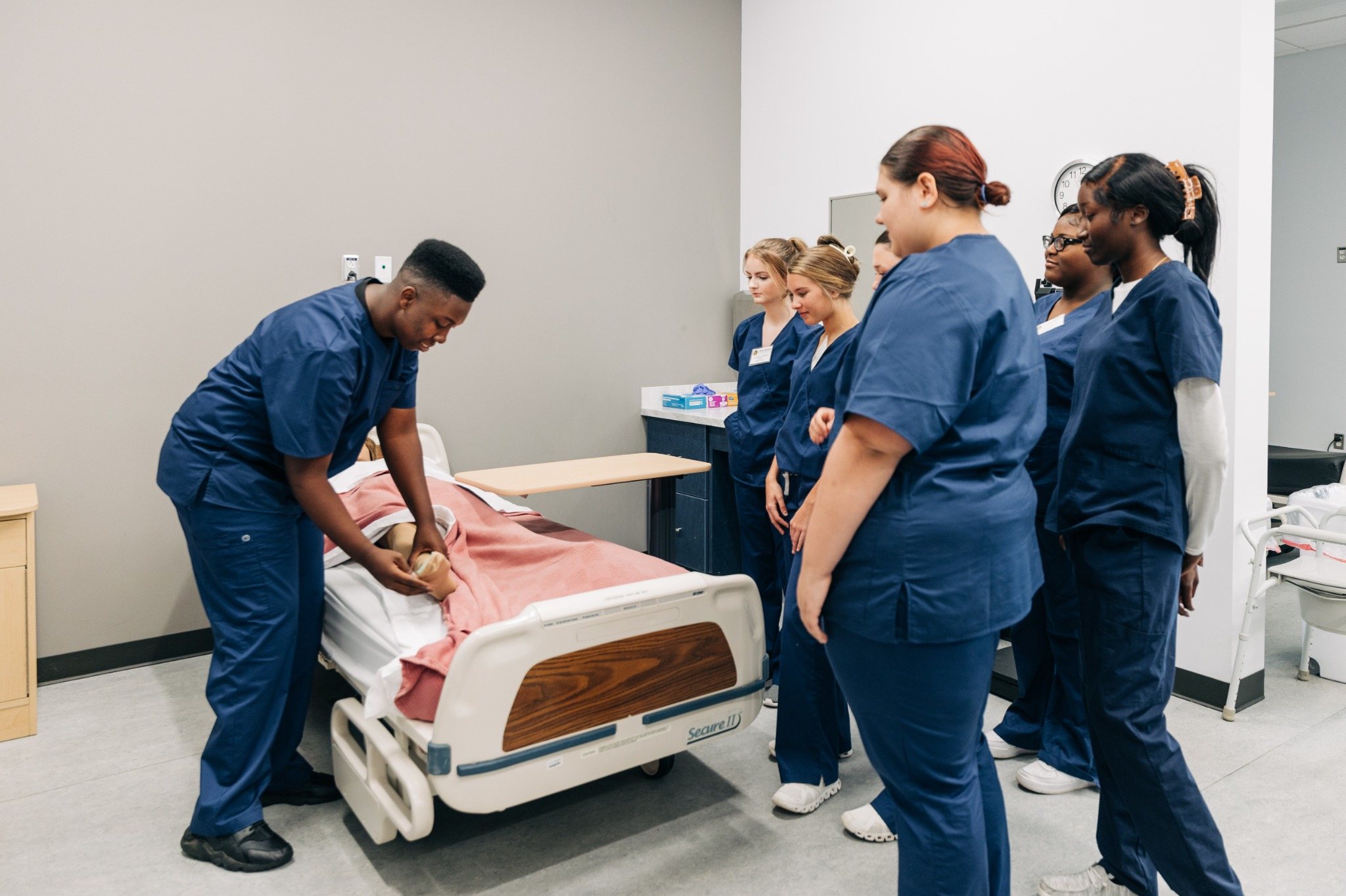 CNA students in blue scrubs observe a classmate practicing patient care skills on a training mannequin at The Continuum.