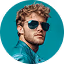 Man with curly hair wearing sunglasses and a blue shirt against a teal background.