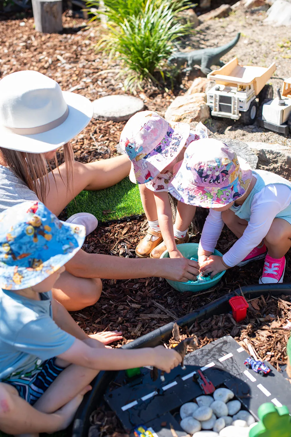 Two toddlers in colorful hats play with water in a blue bowl with an adult nearby outside.