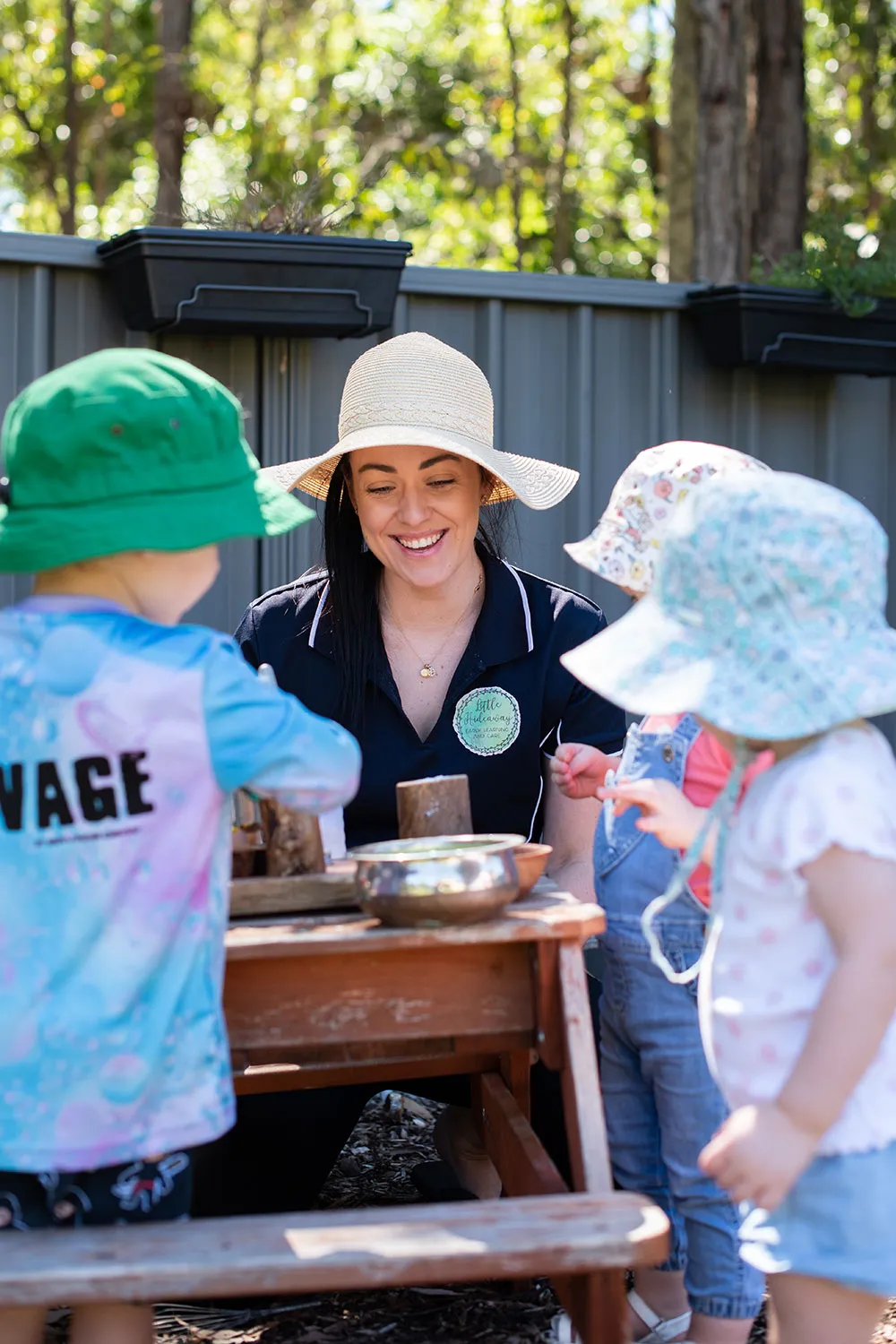 Woman wearing sun hat smiles as three toddlers in hats play around a wooden table outdoors.