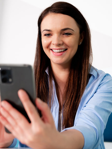 Young woman with brown hair smiling and holding a smartphone in her hand.