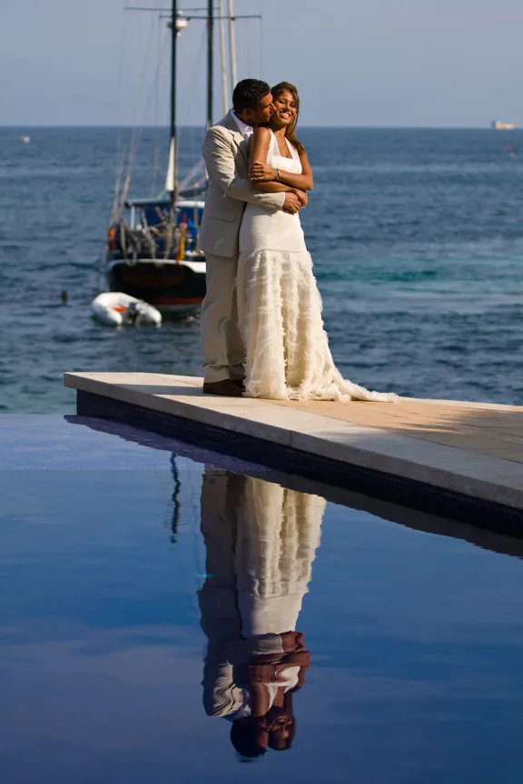 Couple in wedding attire embracing on a poolside platform with ocean and sailboat in the background, their reflection visible in the water.