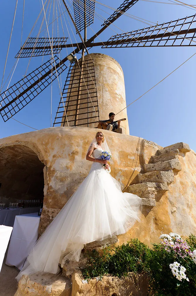 Bride in a flowing white wedding dress holding a bouquet standing on stone steps of a rustic windmill with a man playing guitar seated above.