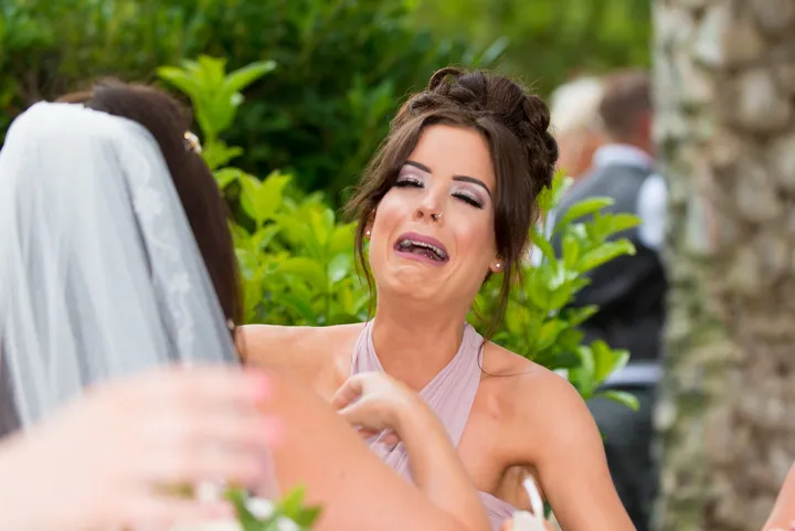 Woman in a light pink dress with an emotional, teary expression hugging a bride wearing a veil outdoors.