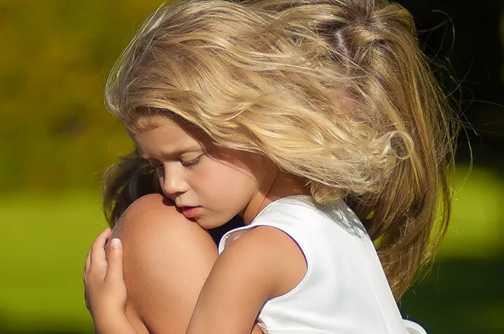 Blonde girl with wavy hair hugging another person outdoors in bright sunlight.
