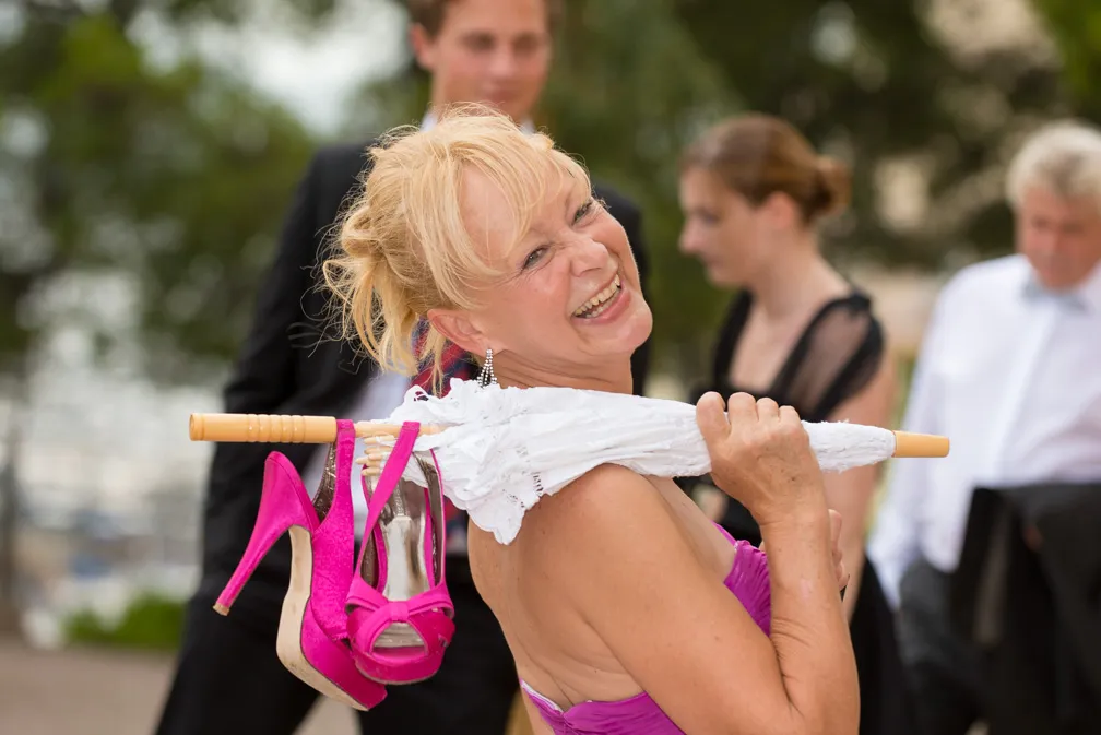Smiling blonde woman in a pink dress holding a white umbrella with pink high heel shoes hanging from it, with blurred people in the background.