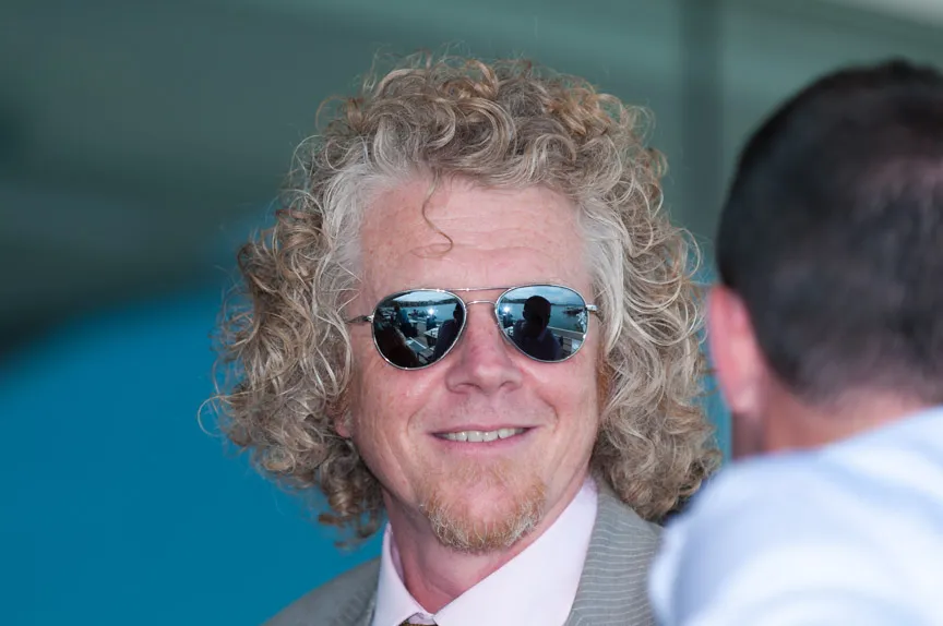 Man with curly blond hair and goatee wearing sunglasses and a suit jacket smiling at another person.