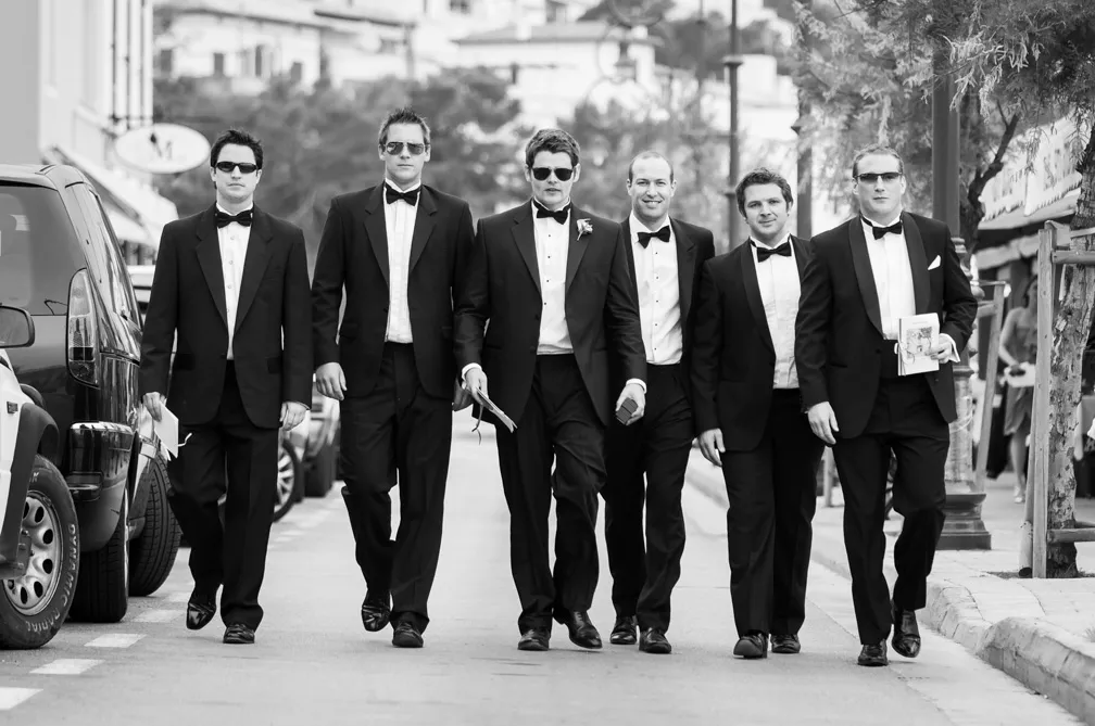 Six men in tuxedos and bow ties walking confidently down a street, with two wearing sunglasses.
