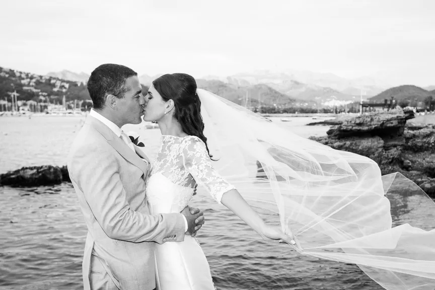 Bride and groom kissing by the water with mountains and boats in the background, bride's veil blowing in the wind.