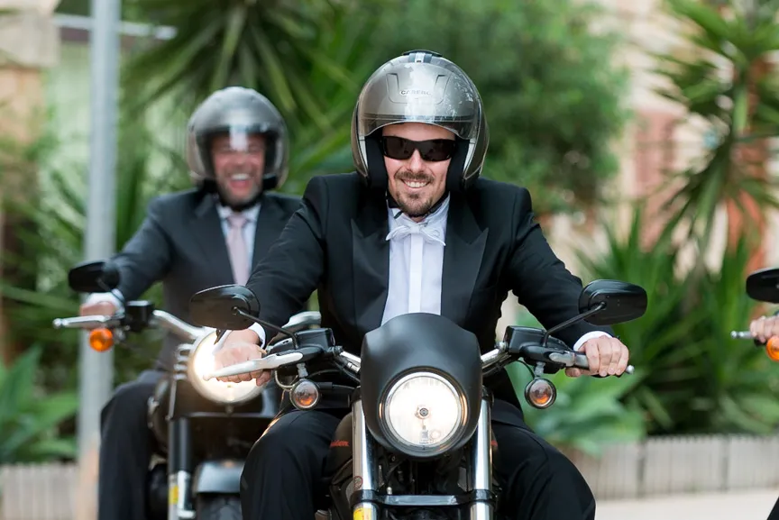Two men wearing helmets and tuxedos riding motorcycles on a street with greenery in the background.