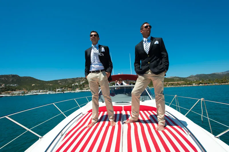 Two men in blazers, striped ties, and khaki pants standing barefoot on the bow of a yacht with a red and white striped cushion under clear blue sky.