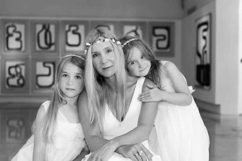 Black and white photo of a woman with two young girls, all wearing white dresses and floral headbands, posing indoors in front of abstract artwork.