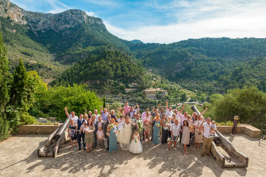 Large group of wedding guests and bride posing happily outdoors with green forested mountains in the background.