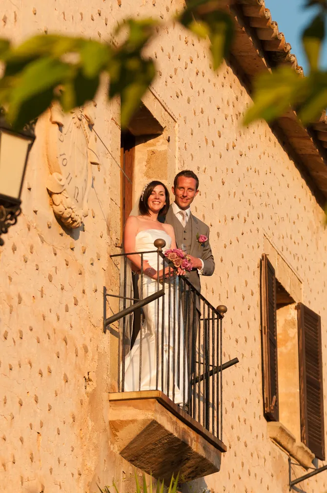 Bride and groom standing on a small balcony of a rustic stone building, smiling and holding a bouquet of roses.
