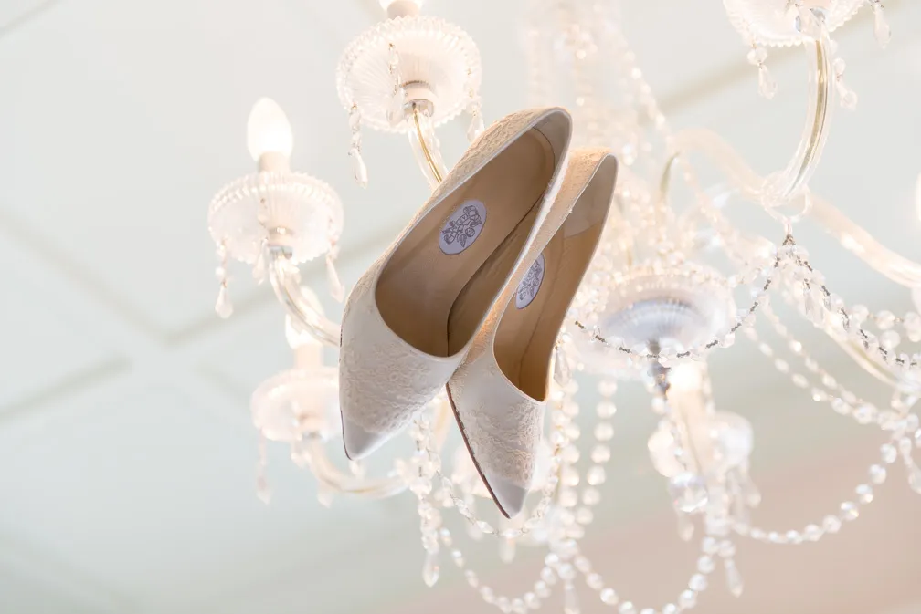 Pair of white lace bridal shoes hanging from a crystal chandelier with lit bulbs.
