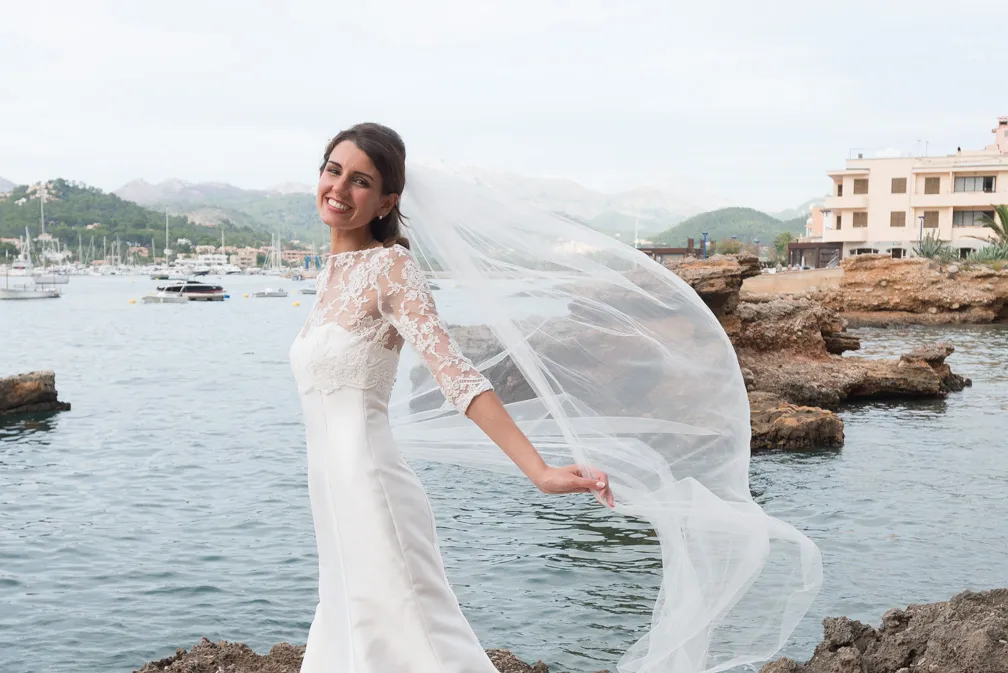 Smiling bride in a white lace wedding dress holding her flowing veil with a coastal background of water, boats, rocks, and buildings.