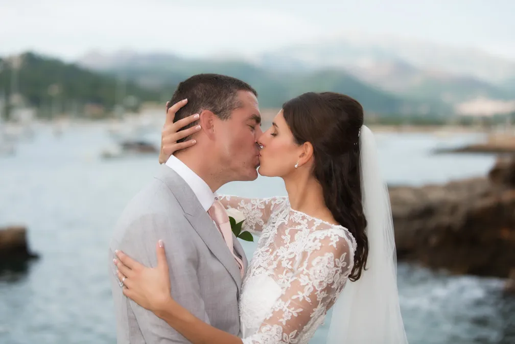 Bride and groom kissing by the water with mountains in the background.