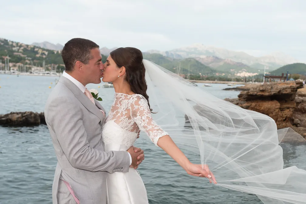 Bride and groom kissing by the waterfront with the bride's veil flowing in the wind and mountains in the background.