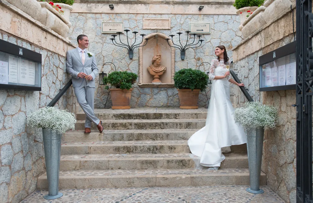 Bride in a white wedding dress and groom in a light gray suit standing on opposite sides of stone steps outside, smiling at each other.