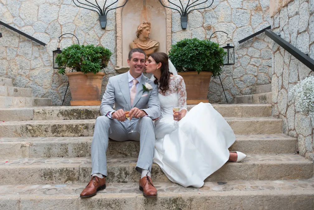 Bride in white gown kissing groom in light gray suit while sitting on stone stairs holding champagne glasses.