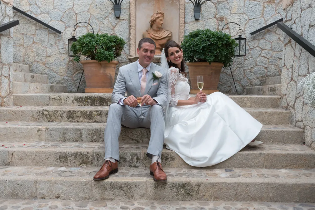 Bride in white dress and groom in light gray suit sitting on stone steps holding champagne glasses.