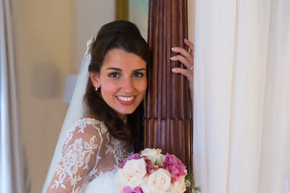 Smiling bride in a lace wedding dress holding a bouquet of white and pink roses, standing beside a wooden pillar with curtains in the background.