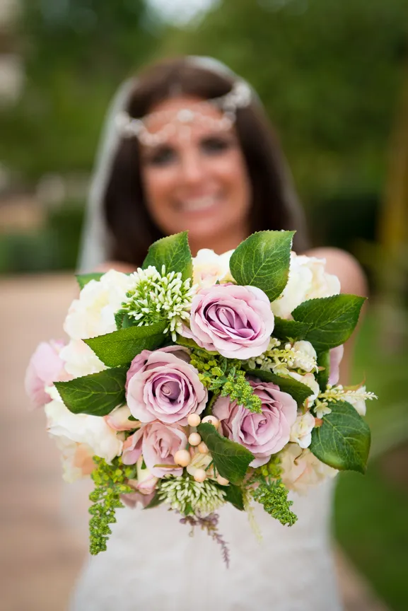 Close-up of a bride holding a bouquet of light pink roses, white flowers, and green leaves with the bride blurred in the background.