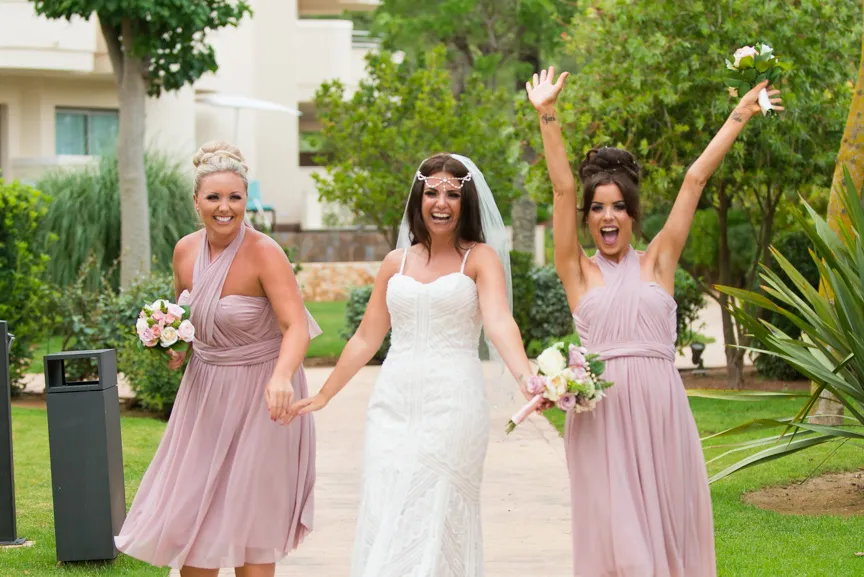 Bride in white wedding dress holding hands with two bridesmaids in pink dresses outdoors, all smiling and joyful.