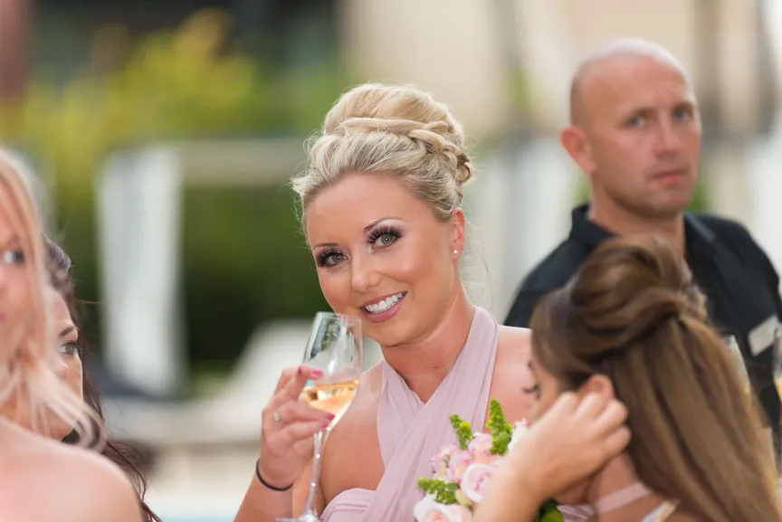 Smiling woman in a pink dress holding a glass of wine and a bouquet, surrounded by people at an outdoor event.