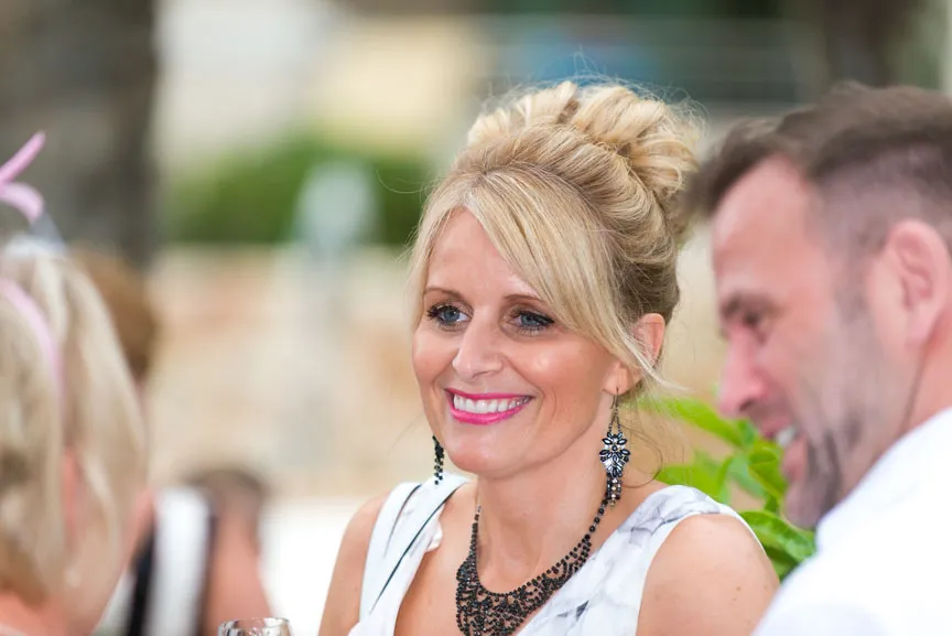 Smiling blonde woman with updo hairstyle wearing black statement necklace and earrings, engaging with two people.