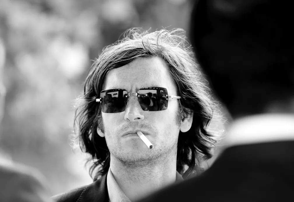 Black-and-white close-up of a man with medium-length hair wearing sunglasses and holding a cigarette in his mouth.