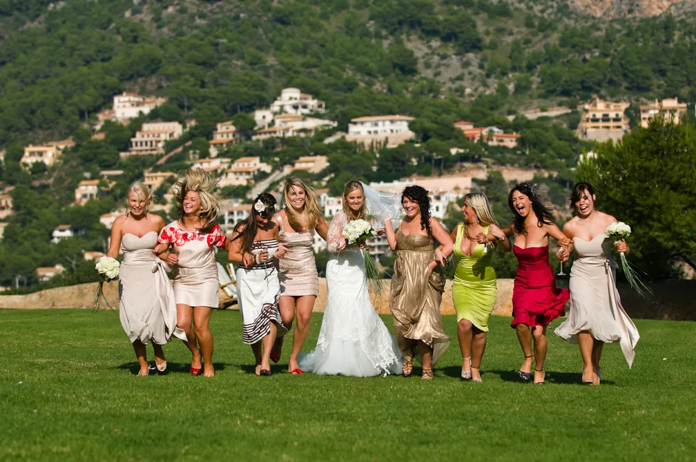 Bride in white dress and bridesmaids in colorful dresses joyfully running together on green grass with hillside houses in the background.