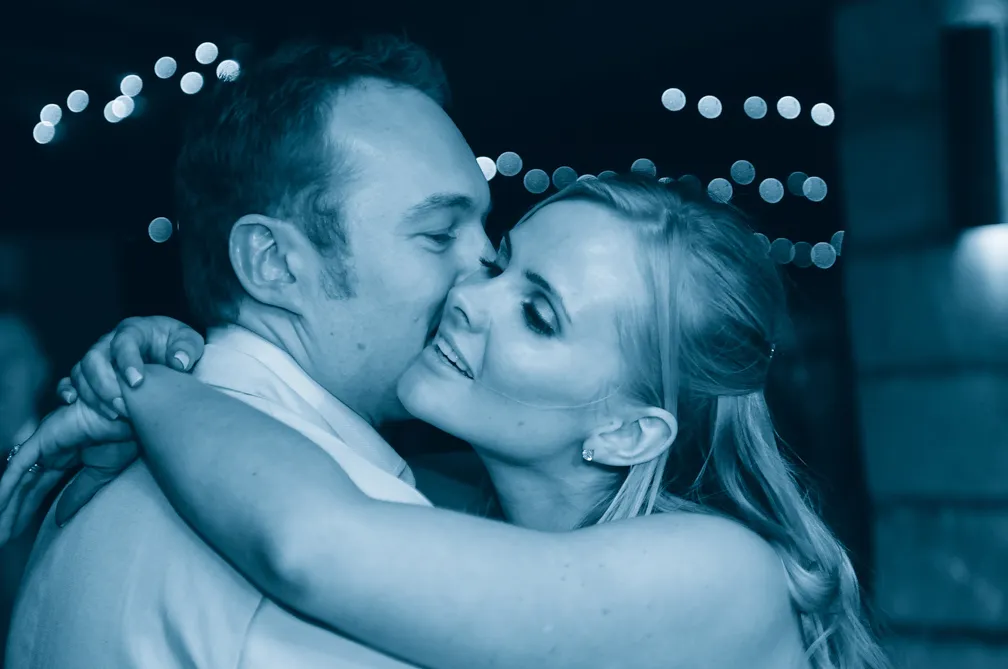 Close-up of a couple embracing closely with the man gently kissing the woman's cheek, against a dark background with blurred string lights.