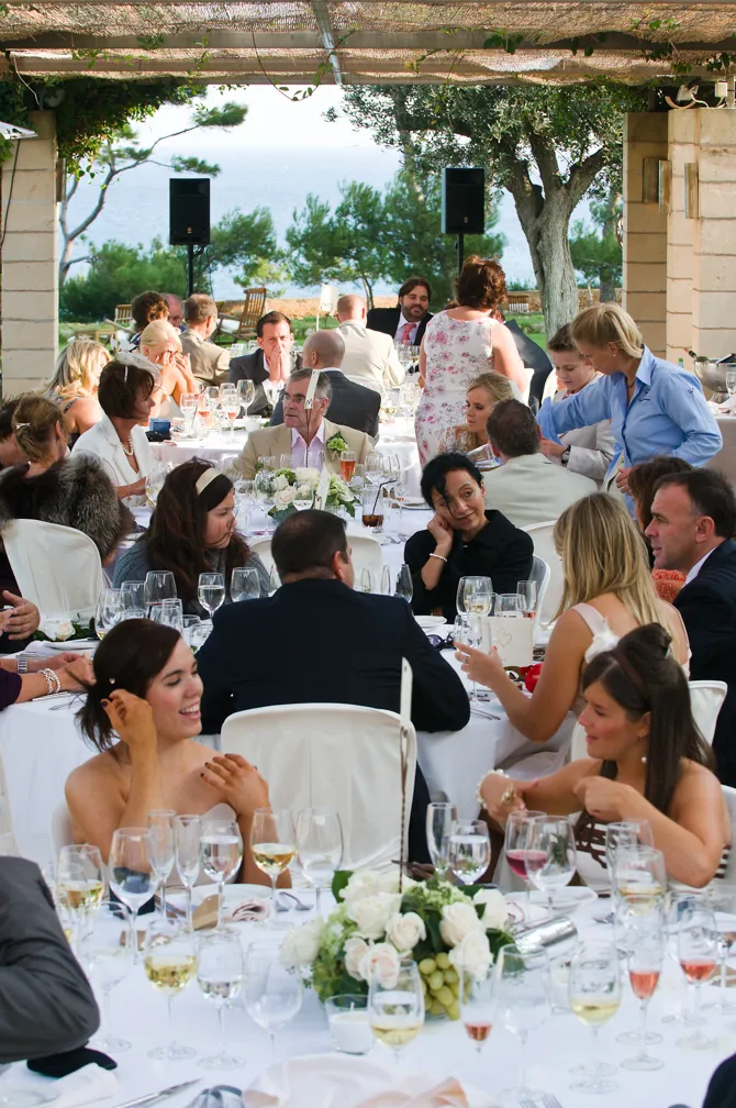 Guests socialize and dine at a formal outdoor event with white tablecloths, floral centerpieces, and a view of trees and water in the background.