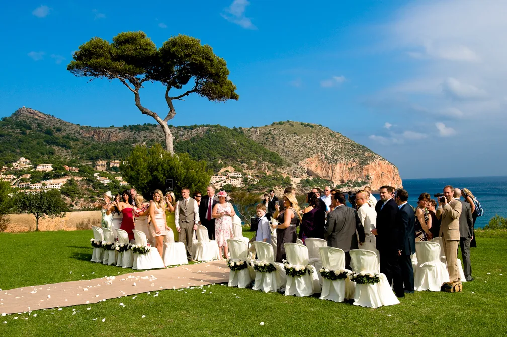 Outdoor wedding ceremony setup with white chairs and guests gathered on a grassy hill overlooking a coastal landscape with hills and the ocean under a clear blue sky.