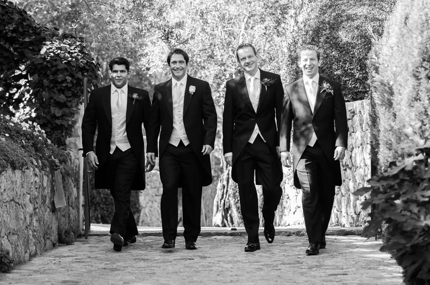 Four men in formal tuxedos walking side by side down a stone pathway outdoors, smiling and looking ahead.