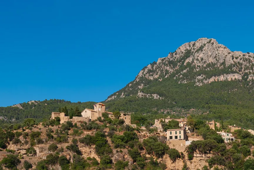 Stone buildings and a church on a hillside covered with trees, with a rocky mountain and clear blue sky in the background.