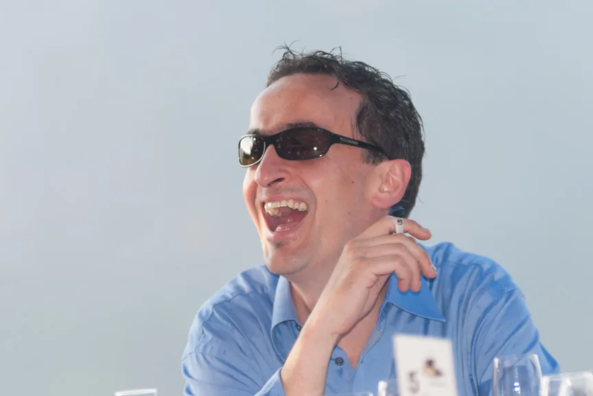 Man wearing sunglasses and a blue shirt laughing and holding a cigarette.