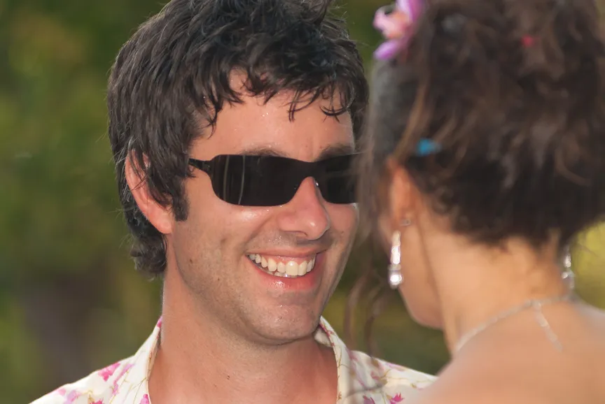 Smiling man wearing sunglasses facing a woman with earrings and a necklace.