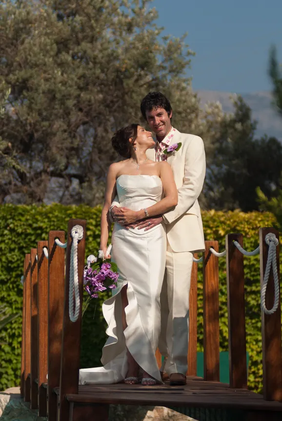 Bride in strapless white gown holding purple flowers embraced by groom in beige suit on wooden bridge outdoors.