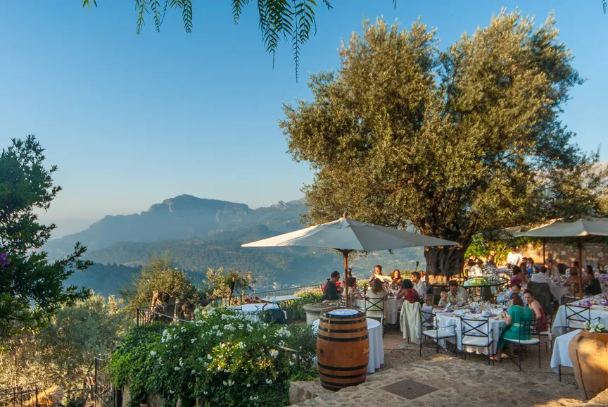 Outdoor dining area with tables, people seated under large umbrellas, greenery, and a scenic mountain view in the background on a clear day.
