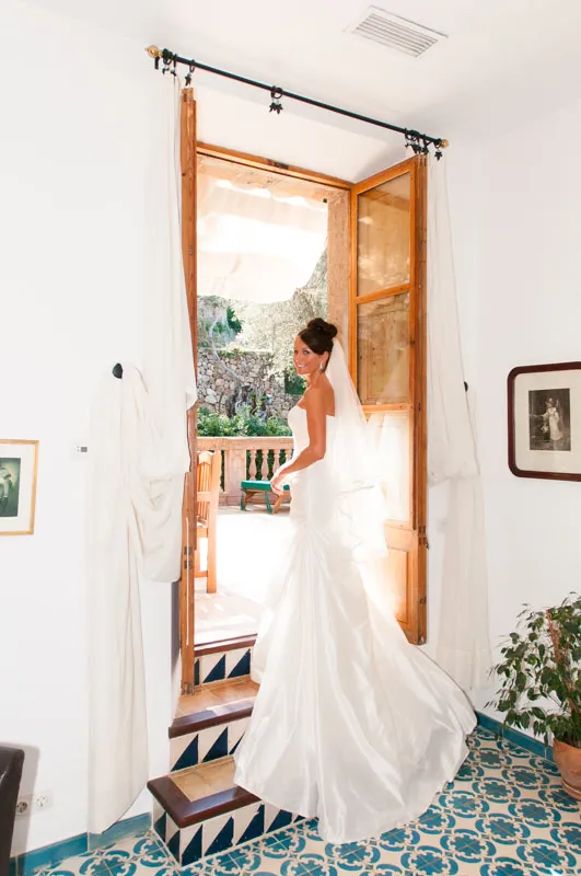 Bride in a white strapless wedding gown standing in a doorway with sunlight streaming in from outside.