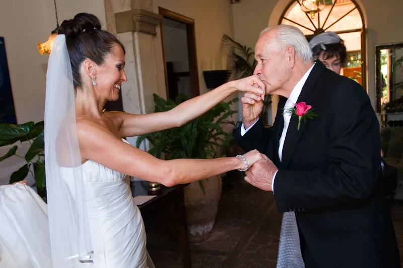 Bride in a white strapless gown and veil smiling as an elderly man in a black suit kisses her hand indoors.