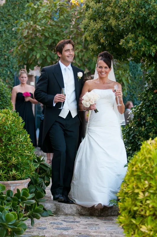 Smiling bride in white gown and groom in black tuxedo walking outdoors, each holding a champagne glass.