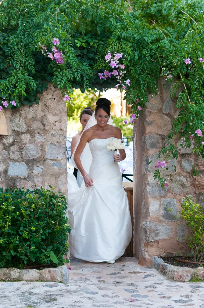 Bride in a white strapless wedding gown holding a bouquet of white flowers, walking through a stone archway decorated with green vines and pink flowers.